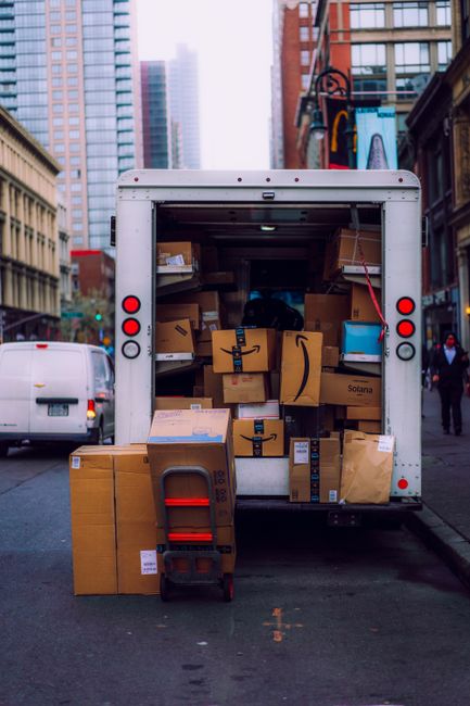 A close-up view of multiple stacked cardboard boxes prepared for home relocation inside a vehicle or loading area, with some boxes displaying 'Fragile' stickers and handling instructions. The boxes ar