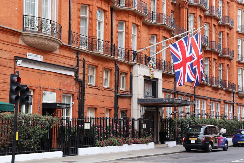 The exterior of a multi-storey red-brick residential building in an urban area, featuring multiple black iron balconies and white-framed windows. Two large Union Jack flags are mounted on the building