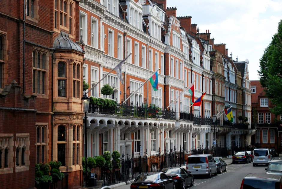 Photograph of a row of elegant, historic terraced houses in Berkeley Square, Mayfair, with red-brick facades, white decorative trim, and large windows. Several flags are mounted on the black wrought-iron balconies, which are adorned with potted plants and flowers. The street features parked cars along the pavement, which is lined with trees providing greenery on the right side. The image captures the exterior architecture and urban environment typical for Mayfair, illustrating the setting for house removals and furniture transport services, with lighting indicating a clear day. Mayfair Movers may utilize such locations for efficient home relocation and packing and moving operations in central London.